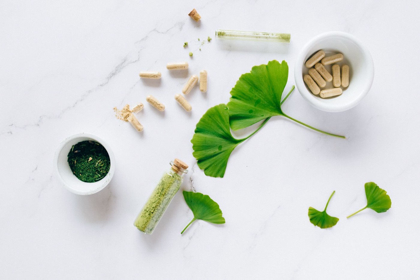Herbal supplement capsules, loose powder and fresh leaves arranged on a marble surface